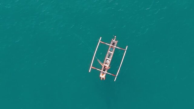 Drone footage of a Balinese fisherman casting a fishing net from a traditional outrigger jukung boat on turquoise ocean water. Overhead view showing the action of fishing at sea