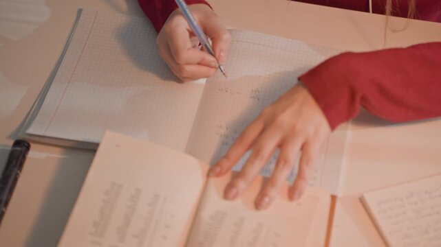 Person in red long sleeve garment writes mathematical equations. Desk includes open books additional notebooks filled. Soft lighting illuminates workspace highlighting study
