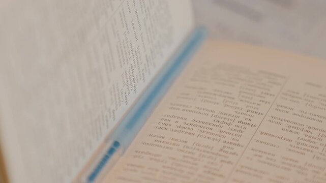 Close up of open dictionary with bilingual text. Camera slowly zooms in and pans across detailed word entries. Soft lighting emphasizes paper texture and text clarity