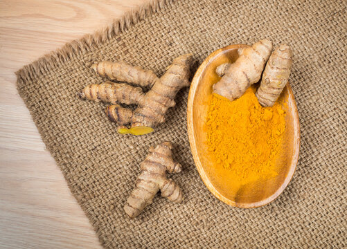 Fresh turmeric roots and turmeric powder in wooden bowl on burlap cloth background top view.