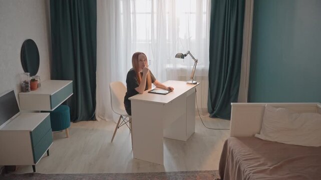 Young woman with long hair sits at white desk in her bedroom. Woman occasionally pauses to think or adjust posture. Room features light colored wooden flooring teal curtains