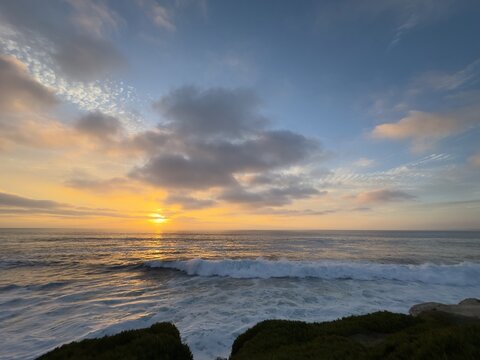 Luminous sunset sky with scattered clouds over crashing ocean waves along the coastal shoreline in La Jolla, California.