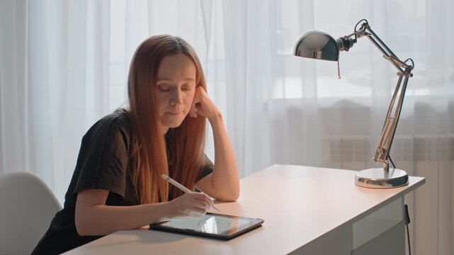 Young woman with long hair draws geometric shapes. Woman uses electronic stylus resting her head. Metallic adjustable lamp sits to her right maintaining consistent