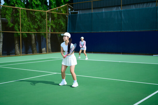 Young Asian Women Playing Tennis Doubles on Outdoor Green Court in Sunny Daylight