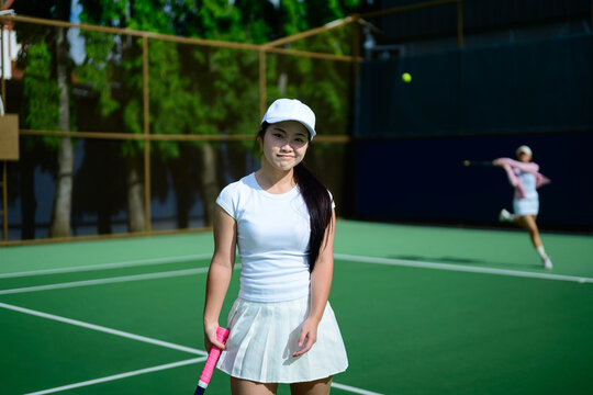 Young Asian Woman Tennis Player Smiling on Outdoor Green Court with Partner Playing in Background