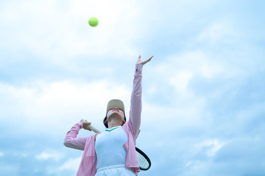 Young Asian Woman Tennis Player Serving Ball Outdoors Against Cloudy Sky, Low Angle Action Shot