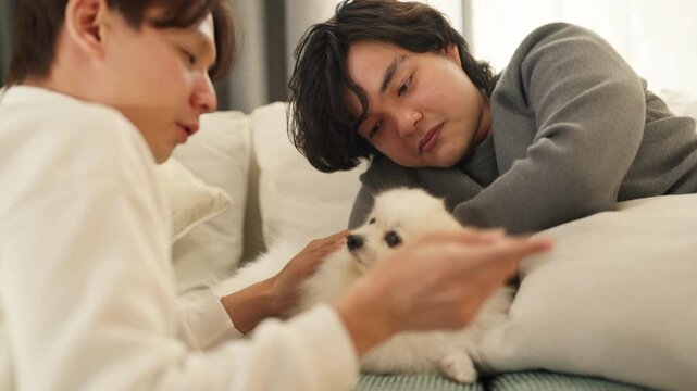 Two smiling friends feeding treat to fluffy Pomeranian puppy on sofa at home, playful moment with dog, pet ownership and companionship concept, cozy living room, natural indoor light.