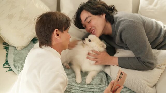 Two smiling friends feeding treat to fluffy Pomeranian puppy on sofa at home, playful moment with dog, pet ownership and companionship concept, cozy living room, natural indoor light.