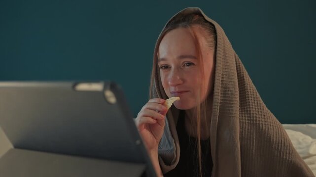 Young woman sits in front of teal wall covered by brown blanket. Woman holds food near mouth with minimal movement. White bedding visible behind her dark electronic device tablet