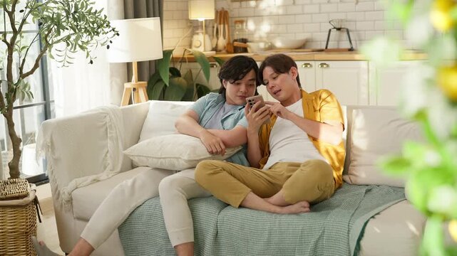 Two male friends relaxing together on sofa at home, one checking smartphone while other leans in closely, cozy living room, casual friendship and bonding lifestyle concept, warm natural light interior