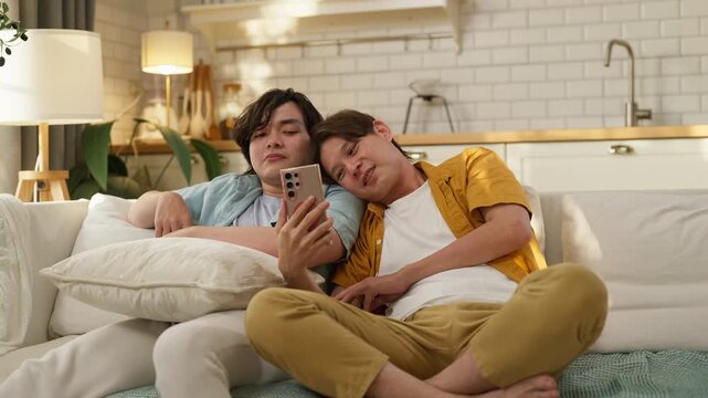 Two male friends relaxing together on sofa at home, one checking smartphone while other leans in closely, cozy living room, casual friendship and bonding lifestyle concept, warm natural light interior