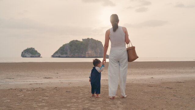 Asian mother holding hands with toddler son walking on sandy beach together with sunset.