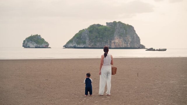 Asian mother holding hands with toddler son walking on sandy beach together with sunset.