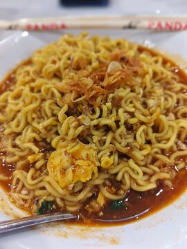 bowl of savory instant noodles (Mie Tek-Tek or Mie Nyemek) served in a thick, spicy, reddish-brown broth.