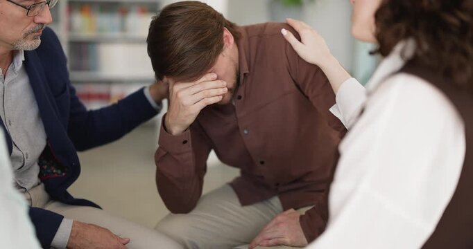 Compassionate groupmates providing psychological support to male participant in emotional distress consoling grieving peer on group therapy session offering reassurance encouragement understanding