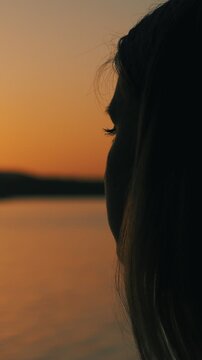 Close-up of a girl's face admiring the sunset at dusk on the shore of a lake. Portrait of a woman at sunset. Human inspiration. Vertical video.