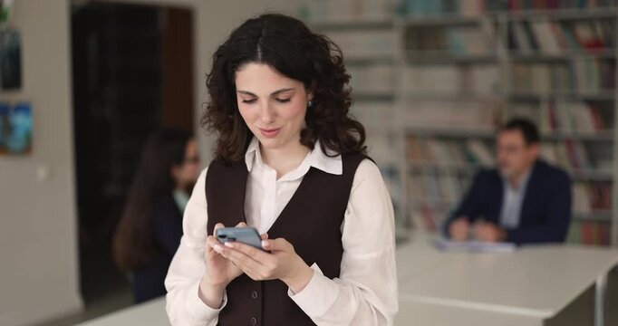Busy young adult female college or university student messaging on mobile phone in campus library browsing educational resources searching literature in electronic catalog checking lecture schedule