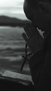 A religious young man reads the Bible and prays to the Lord God on the shore of a lake. He holds a Christian cross in his hands. Black and white. Vertical video.