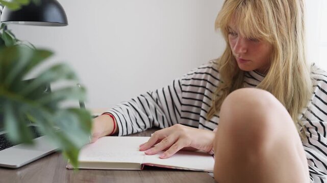 Creative girl sitting at a desk near a bright window. Concentrating while writing or drawing in her notebook with a pencil. Showcasing a moment of focused study or creativity