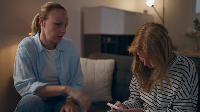 Mother talking to teenage daughter at home while girl ignores her and focuses on smartphone, illustrating excessive screen time, digital distraction, parenting challenges and family communication.