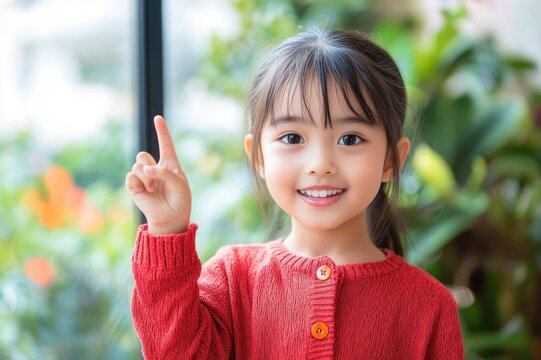 young child in red sweater pointing upward by a window with green plants, curious thoughtful gesture