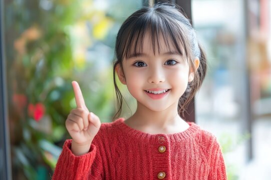 young girl in red knit sweater raising one finger by a window with blurred green plants, thoughtful and curious expression
