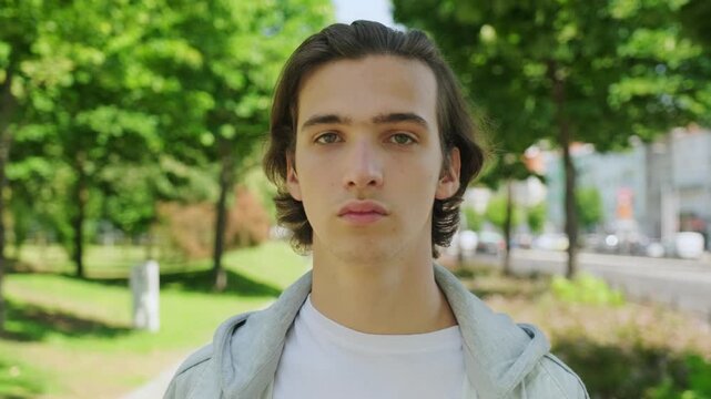 Serious young  man walks and  looks in the camera, outdoors. Calm and peace look of an young man.  Closeup  calm face of  handsome guy at the park.  White  male person face, in the city. Slow motion