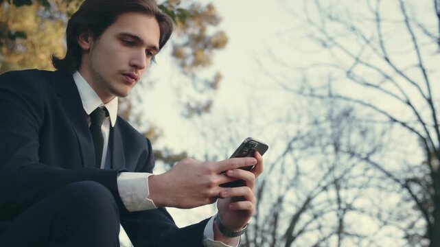 Confident man in a business suit is sitting outdoors and using  smartphone. A young man in a suit with mobile phone, slow motion. Serious and calm businessman in the city  looking to a cell phone.