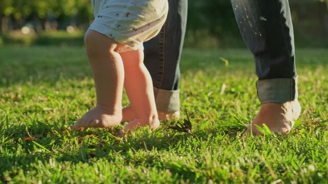 Walking children's bare feet on a green lawn close-up. Child  learns to take the first steps.  Baby learns to walk with the help of his mother on a green grass in the park. Slow motion 4k video.