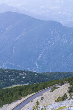 A narrow paved road descends through rocky terrain and scattered pine trees on a high mountainside overlooking a hazy patchwork valley below. Distant hills fade into soft blue atmospheric haze