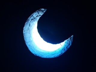 Crescent moon shaped hole in a rough stone or concrete wall with bright light shining through from behind against a dark background. © LoneFighter