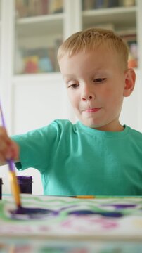 Vertical lifestyle video of a happy child learning to draw with a brush and paint in kindergarten. Kid painting with colorful watercolors at his desk.