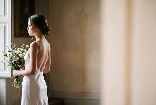 Young woman in white dress holding bouquet by window