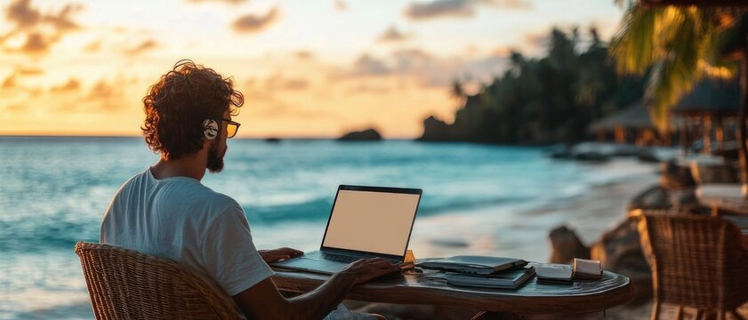 curly-haired person with headphones and glasses working on a laptop at a wicker table on a tropical beach at sunset with notebooks and phone nearby, calm and focused mood