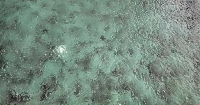Slow motion close-up of raindrops creating rhythmic ripples on the surface of a swimming pool, revealing the tiled bottom.