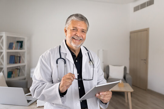 Smiling senior male doctor, holding digital tablet in medical clinic office. Healthcare industry, online consultation using telemedicine application, and professional general practitioner portrait