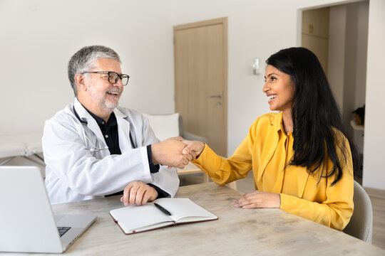 Smiling male physician doctor sitting at desk, greeting female patient with friendly handshake, starting medical clinic consultation, providing professional services. Trust and agreement in healthcare