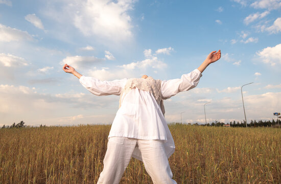Low-angle shot of a person dressed in white clothing and a knit vest, standing in a golden wheat field with arms outstretched toward a blue sky with white clouds. Young woman dancing outdoors