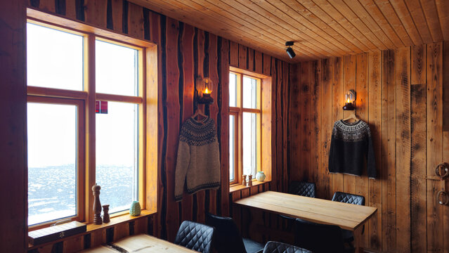 Interior view of a cozy, wood-paneled cabin or restaurant in Iceland featuring rustic wood walls, large windows overlooking a bright landscape, traditional woolen sweaters hanging on the walls, and a 