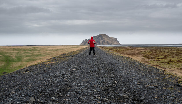 A rear-view shot of a lone traveler wearing a bright red jacket standing in the middle of a gravel path leading toward a distinct hill under an overcast sky in Iceland.. Adventure Exploration