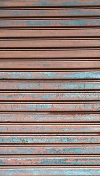 Close-up of an old, weathered metallic rolling shutter door with peeling rust-orange and faded teal-blue paint. The repetitive horizontal corrugated lines show a textured pattern of age and decay.