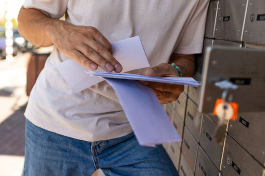 A young man stands at a mailbox with envelopes and reads a letter. Mail recipient. A man stands on the street and holds envelopes in his hands. Close-up of his hands.
