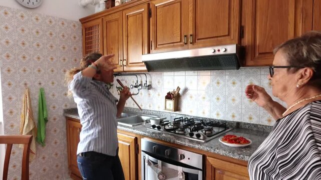 Joyful daughter and senior mother dancing and singing while cooking together in the kitchen. Happy family moments, intergenerational bonding, and positive domestic lifestyle concept