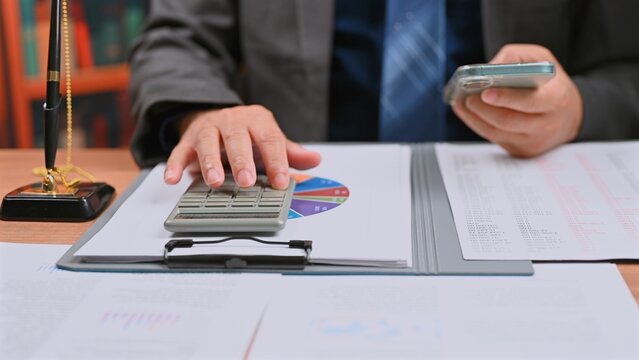 Smart executive does accounting work, reviewing accounting files prior tax audit, as accounting staff finishes audit tasks, pressing key calculator buttons near desk top.