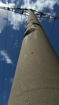 Concrete utility pole damaged by shrapnel from a shell blast, exposed rusted rebar and cracks under overhead power wires. War damage to electricity infrastructure and blackout risk