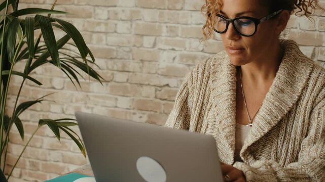 Serious adult woman with glasses concentrating on her work, typing on a laptop keyboard at her home office, embodying the concept of remote work, freelancing, and digital nomad lifestyle
