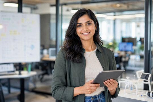 Confident Indian businesswoman holding digital tablet in modern office. Professional female entrepreneur in creative workspace. Successful young leader.