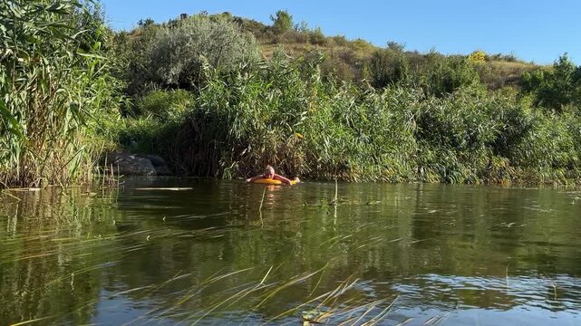 Woman floats on a yellow inflatable ring in a calm green river beside tall reed beds, with willows and a grassy hill under clear blue sky on a late summer day