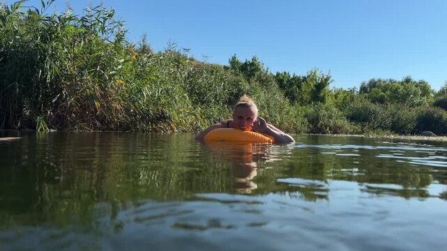 Smiling mature woman swims on an orange inflatable ring in a calm river, waving and spreading her arms wide beside reed beds under clear blue sky in late summer