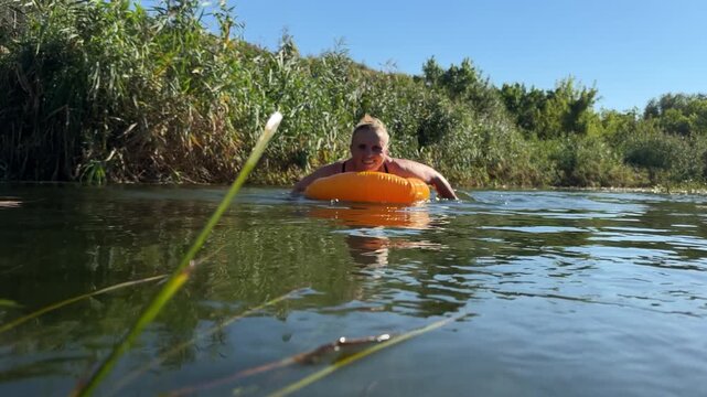 Laughing mature woman swims toward camera on an orange inflatable ring in a calm river, spreading her arms and raising wet palms to lens on a sunny late summer day